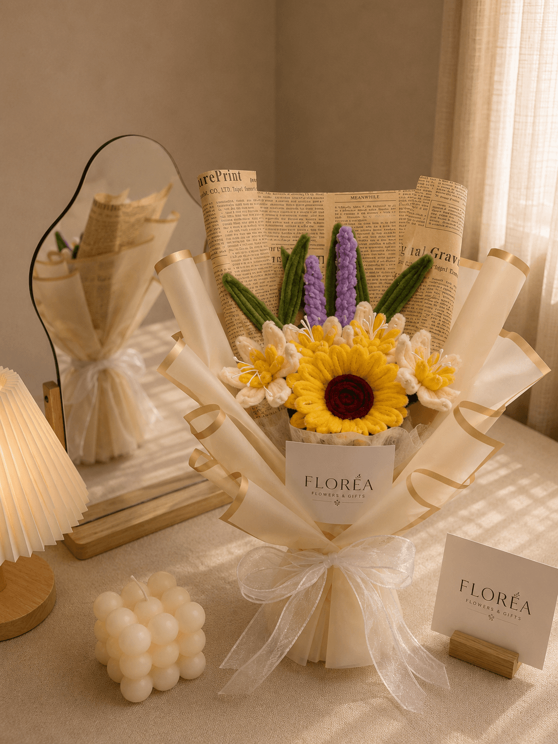 Handmade fuzzy-wire bouquet with a yellow sunflower and red rose center, purple lavender stems, and cream lily accents, wrapped in kraft newspaper with a sheer ribbon, styled on a sunlit tabletop with a mirror