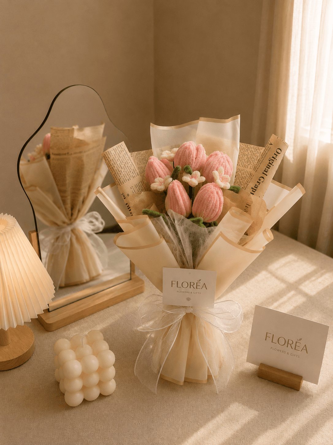 Handmade fuzzy-wire pink tulip bouquet with white filler flowers, wrapped in kraft newspaper and ivory paper with a sheer white ribbon, styled on a sunlit tabletop with a mirror